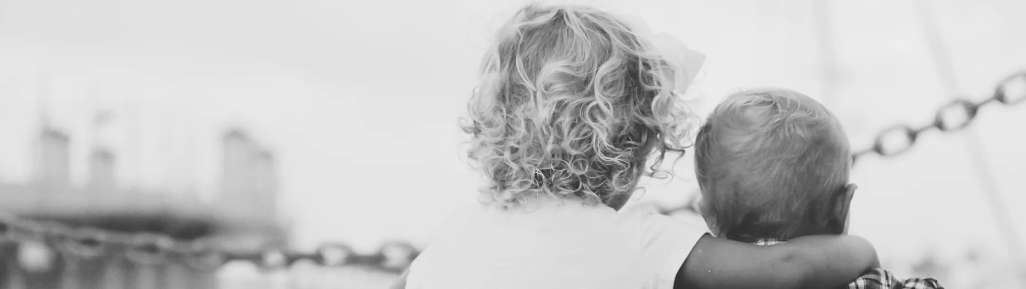 Black-and-white photo of two small children seen from behind, sitting side by side at the water’s edge, one with an arm around the other. Photo: Joshua Clay, Unsplash via Wikimedia Commons — CC0 (public domain).